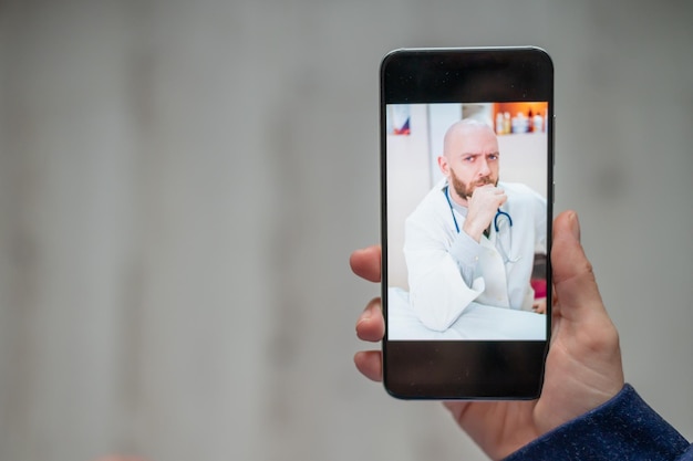 Gros plan sur des mains féminines avec un téléphone Un médecin sérieux, chauve et barbu, mène une consultation en ligne avec le patient via une webcam Une femme parle au médecin traitant par le biais d'un appel vidéo