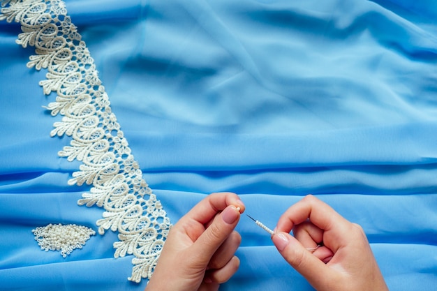 Gros plan sur les mains d'une femme couturière tailleur (couturière) robe en mousseline de soie bleue coud des perles à lacer sur fond bleu dans le studio