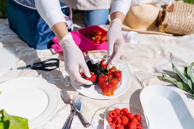 Gros plan sur les mains de la femme avec des gants mettant des fruits rouges dans une assiette. Préparation d'un pique-nique en plein air.