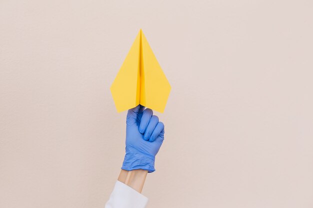 Gros plan sur les mains des femmes dans des gants de protection en latex bleu tenant un avion en papier jaune sur fond beige. Espace de copie