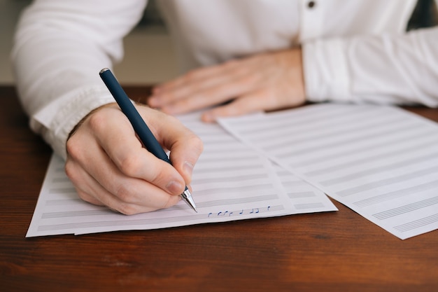 Gros plan des mains d'un homme méconnaissable écrivant des notes de musique avec un stylo sur des partitions