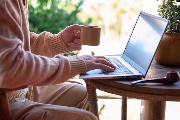 Gros plan sur les mains d'un homme senior naviguant sur un ordinateur portable en plein air assis sur le balcon en tapant sur un clavier tenant une tasse de café Table rustique en bois dans un chalt de montagne