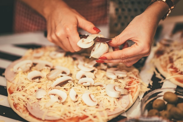 Gros plan sur les mains d'une jeune femme coupant des champignons et mettant la pâte à pizza.