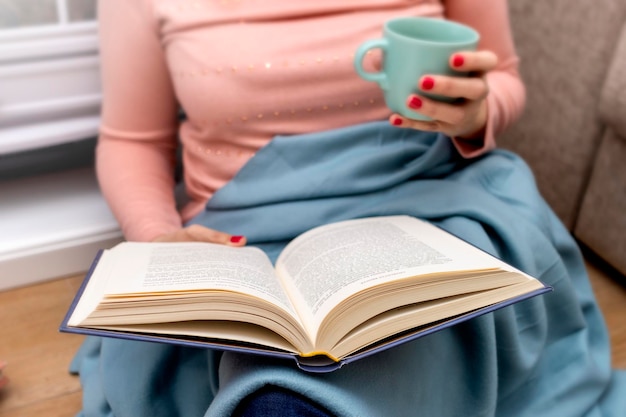 Gros plan sur les mains d'une jeune femme avec une tasse de café lisant un livre bleu à couverture rigide à la maison