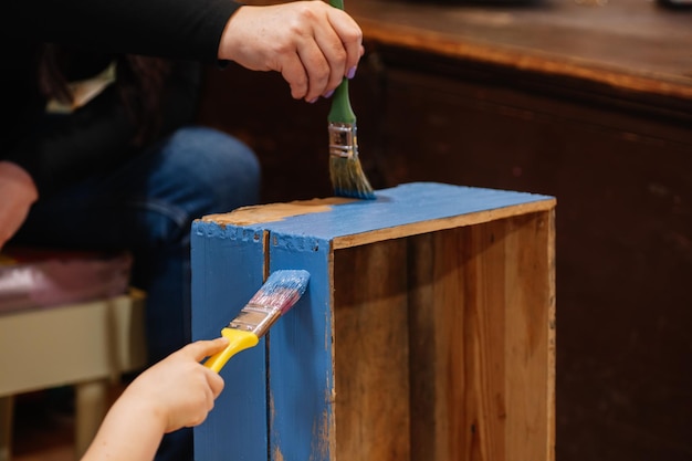 Gros plan sur les mains d'une jeune fille et d'une femme avec des pinceaux peignant lentement un très vieux tiroir de placard en bois de couleur bleue Réutilisation de vieilles choses Actions écologiques durables pour l'avenir de la planète