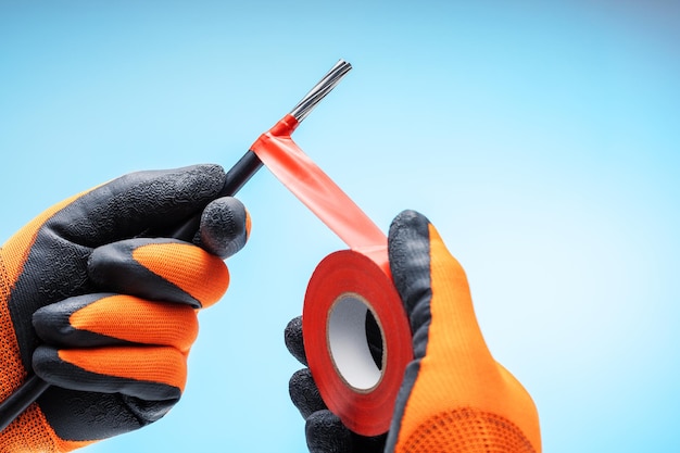 Gros plan des mains mâles dans des gants tenant un rouleau de ruban adhésif rouge pour les travaux électriques Isolation du câble électrique