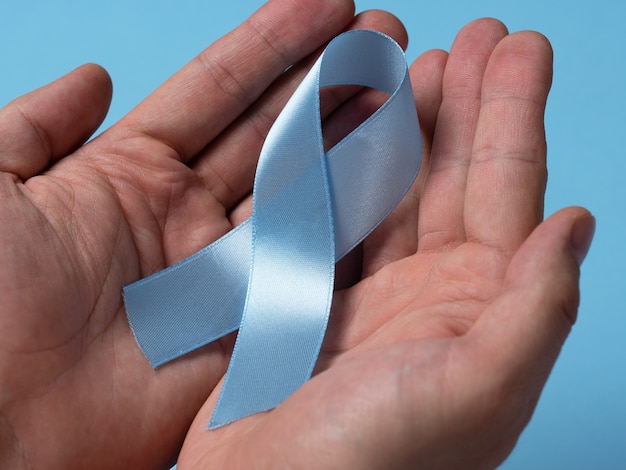 Gros plan des mains masculines tenant un ruban bleu de sensibilisation au cancer de la prostate sur un fond bleu clair. Concept de médecine et de soins de santé