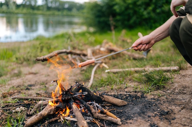 Gros plan des mains d'un randonneur méconnaissable grillant des saucisses sur une cheminée dans la côte de la rivière