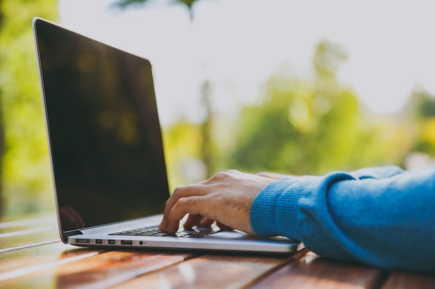 Gros plan des mains recadrées sur le clavier. Homme d'affaires ou étudiant intelligent réussi assis à table avec un téléphone portable dans le parc de la ville à l'aide d'un ordinateur portable, travaillant à l'extérieur. Concept de bureau mobile. Vue de côté.