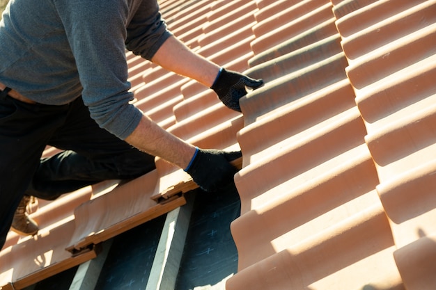 Gros plan des mains des travailleurs installant des tuiles de toiture en céramique jaune montées sur des planches de bois couvrant le toit d'un bâtiment résidentiel en construction.