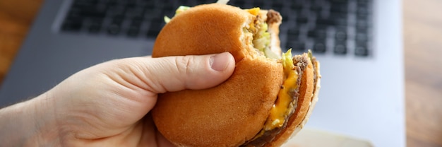 Gros plan, mâle, main, tenue, cheeseburger Travailleur mangeant de la restauration rapide au lieu de travail au bureau. Ordinateur portable et boîte sur table en bois. Malbouffe et concept de pause déjeuner