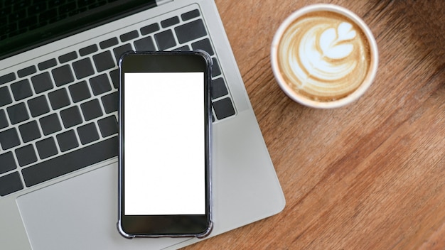 Gros plan d'une maquette d'un smartphone à écran blanc sur clavier d'ordinateur portable avec café latte sur table en bois.