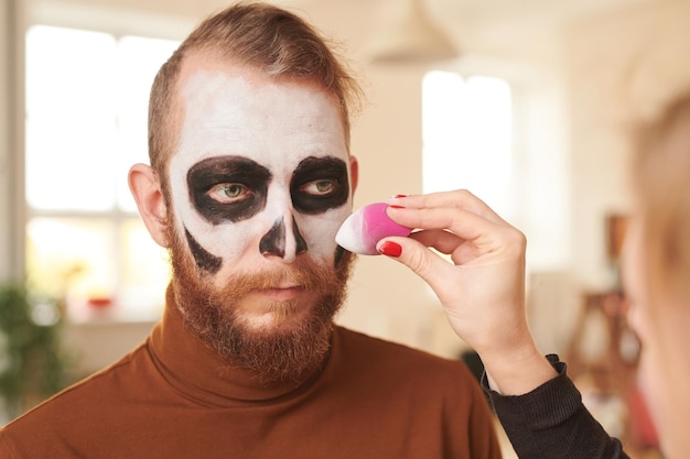 Gros plan d'un maquilleur méconnaissable à l'aide d'une éponge tout en rendant le visage des hommes blancs pour la fête d'halloween