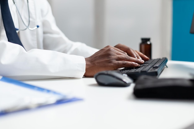 Gros plan d'un médecin afro-américain tapant une ordonnance de médicament sur un ordinateur travaillant au traitement de santé au bureau de l'hôpital. Spécialiste analysant l'expertise de la maladie surveillant les symptômes