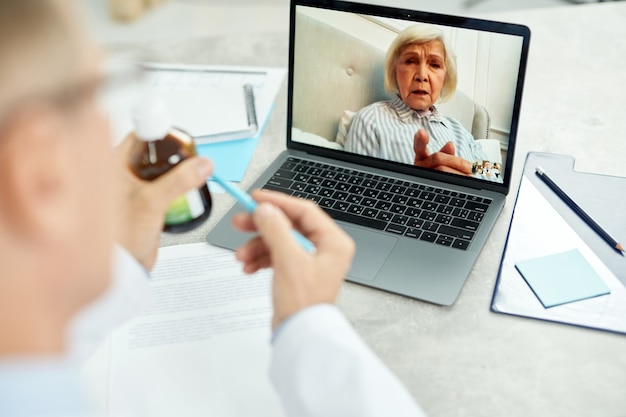 Gros plan sur un médecin de sexe masculin assis à la table avec un ordinateur portable et tenant une bouteille de médicament tout en parlant avec une vieille femme par appel vidéo