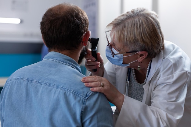 Gros plan sur un médecin tenant un otoscope pour examen de l'oreille avec un jeune homme. Otologue faisant des consultations à l'aide d'un outil d'otologie pour trouver un diagnostic et un traitement de soins de santé dans le cabinet pendant la pandémie.