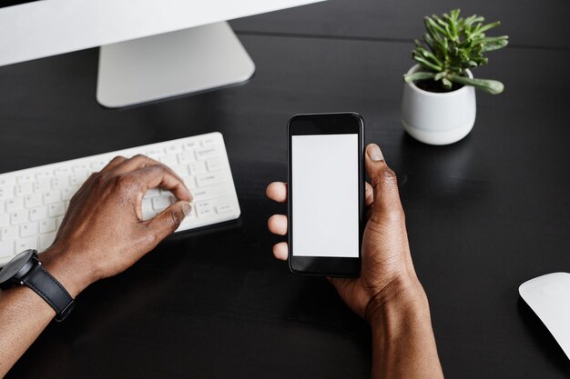 Gros plan minimal de mains masculines tenant un smartphone avec écran vide tout en travaillant sur une copie de bureau noir...