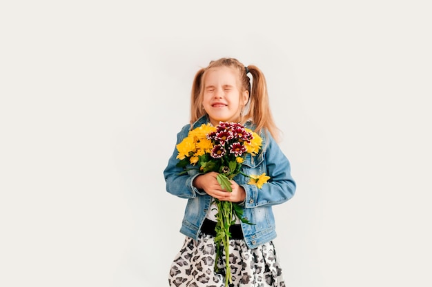 Gros plan, mise au point sélective. Petite fille blonde dans une tient des chrysanthèmes et des gerberas dans ses mains sur fond blanc, une fillette sourit et tient des fleurs de printemps dans ses mains