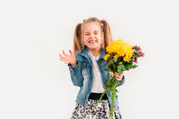 Gros plan, mise au point sélective. Petite fille blonde dans une tient des chrysanthèmes et des gerberas dans ses mains sur fond blanc, une fillette sourit et tient des fleurs de printemps dans ses mains