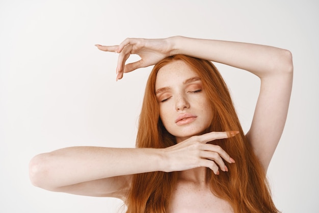 Gros plan d'un modèle féminin rousse tendre avec de longs cheveux naturels rouges, une peau parfaite sans maquillage, montrant un visage propre, debout nu avec les yeux fermés sur un mur blanc