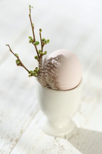 Gros plan d'oeuf de Pâques dans une charrue avec plume de caille et brindille avec bourgeons sur une table shabby chic blanc