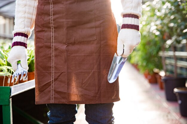 Gros plan des outils de jardin pour la transplantation de plantes tenues par les mains d'un jardinier masculin dans des gants en serre