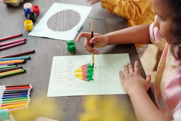 Gros plan sur une petite fille africaine assise à la table et peignant une carte de voeux avec des peintures pour Pâques