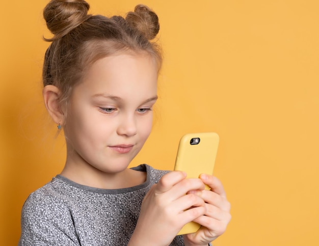 Gros plan d'une petite fille concentrée regardant son smartphone en se tenant debout sur un fond rose. L'enfant aime jouer à des jeux au téléphone. Place pour le texte.