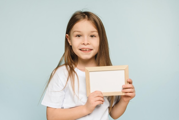 Photo gros plan petite fille tenant un cadre blanc blanc posant isolé sur fond bleu pastel en studio copier la maquette de l'espace