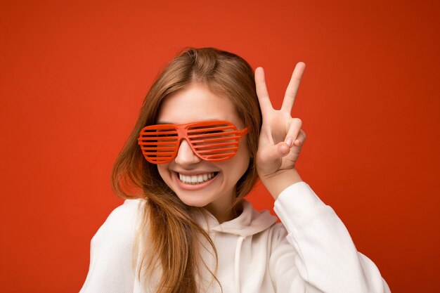 Gros plan photo de charmante jeune femme blonde souriante heureuse isolée sur coloré portant un sweat à capuche blanc décontracté et des lunettes élégantes regardant la caméra et montrant le geste de paix