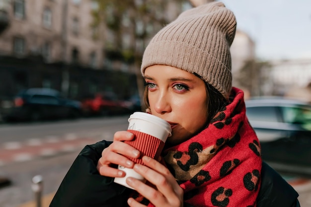 Gros plan photo extérieure d'une belle fille aux yeux verts portant une casquette tricotée et une écharpe rouge appréciant de marcher dans une ville ensoleillée et de boire du café
