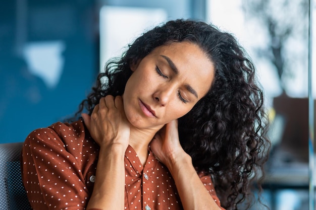 Gros plan photo fatiguée jeune femme hispanique elle est assise dans le bureau tient et masse son cou avec elle