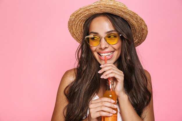 Gros plan photo de femme européenne des années 20 portant des lunettes de soleil et chapeau de paille de boire du jus de bouteille en verre, isolé sur mur rose