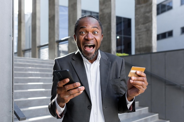 Gros plan photo heureux et choqué homme afro-américain en costume debout à l'extérieur du bureau de la banque, il détient un