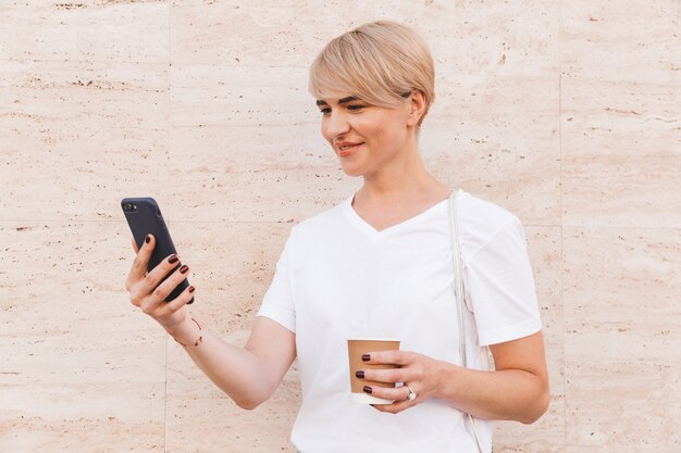 Gros plan photo de jolie femme blonde portant un t-shirt blanc à l'aide de téléphone portable, debout contre un mur beige en plein air en été et tenant une tasse de café en papier