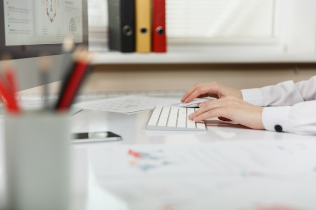 Gros plan photo de mains féminines sur le clavier. Belle femme d'affaires travaillant sur ordinateur avec des documents dans un bureau léger.