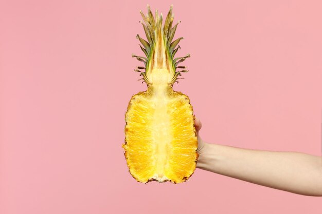 Gros plan sur une photo recadrée d'une femme tenant dans la main un demi-fruit d'ananas mûr frais isolé sur fond de mur pastel rose. Copiez la maquette publicitaire de l'espace. Mode de vie vivant des gens, concept de vacances relaxantes.