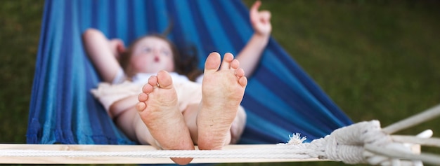 Gros plan des pieds de la petite fille se relaxant dans le hamac bleu pendant ses vacances d'été dans la cour arrière