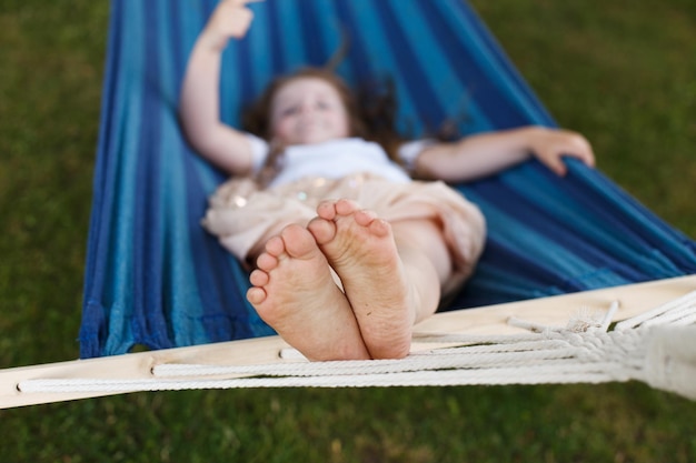 Gros plan des pieds de la petite fille se relaxant dans le hamac bleu pendant ses vacances d'été dans la cour arrière