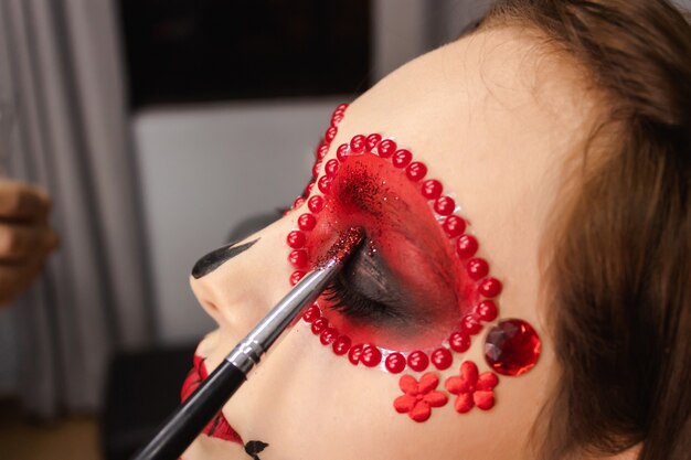 Gros plan sur un pinceau ajoutant des paillettes sur le bord des yeux d'une jeune fille, maquillage Dia de los muertos.