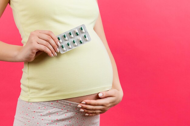 Gros plan d'une plaquette de pilules dans la main de la femme enceinte contre son ventre sur fond coloré avec espace de copie Vitamines pendant la grossesse concept