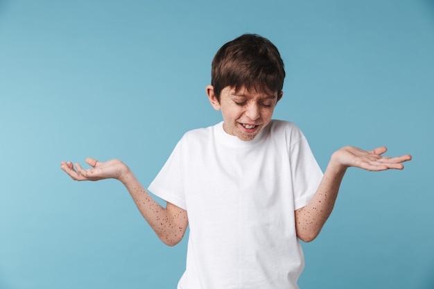 Gros plan Portrait de beau garçon candide avec des taches de rousseur portant un t-shirt décontracté blanc souriant et gesticulant les mains de côté isolé sur mur bleu