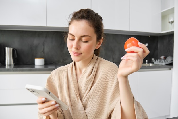 Gros plan portrait d'une belle femme souriante tenant des tomates fraîches assise dans la cuisine avec un smartphone