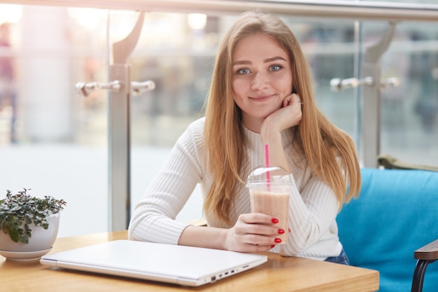 Gros plan le portrait de la belle jeune femme de race blanche aux longs cheveux blonds assis dans un café, boire un cocktail de lait, se reposer après avoir travaillé en ligne sur un ordinateur portable, regardant directement la caméra.