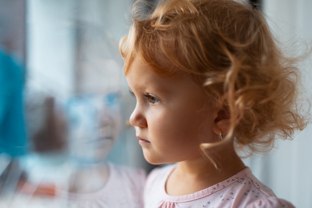 Gros plan portrait de fille triste enfants regardant à travers la fenêtre.