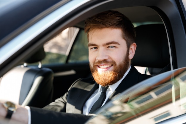 Gros plan le portrait d'un homme barbu souriant en costume au volant de voiture