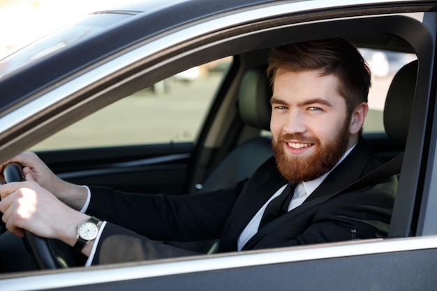 Gros plan le portrait d'un homme barbu souriant en costume au volant de voiture