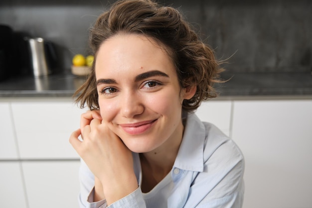 Gros plan, portrait d'une jeune femme brune regarde la caméra et sourit, assise à la maison dans la cuisine