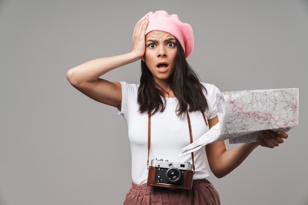 Gros plan portrait d'une jeune femme touristique choquée avec un appareil photo vintage rétro saisissant sa tête tout en tenant une carte papier isolée sur un mur gris