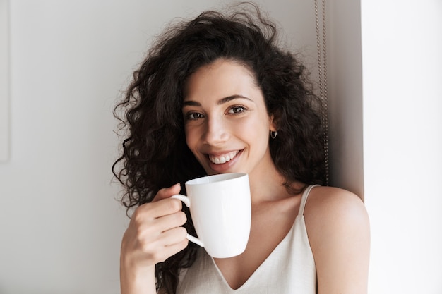 Gros plan Portrait de magnifique femme heureuse avec de longs cheveux bouclés souriant et boire du thé dans une tasse le matin à l'hôtel